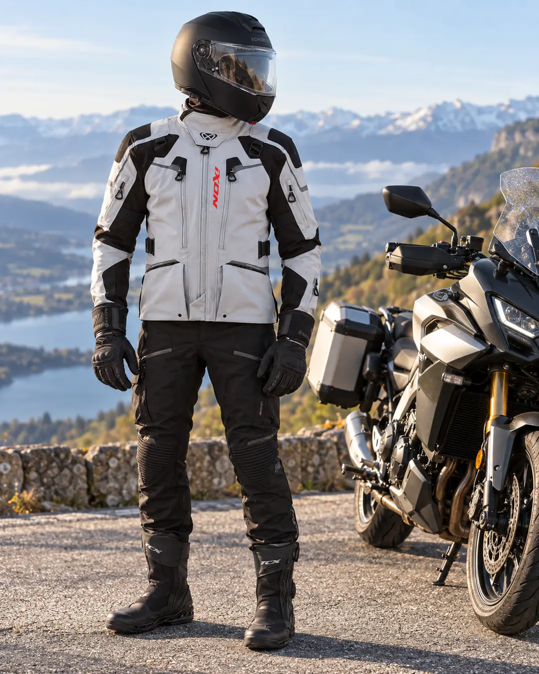 Motard debout près d’un trail routier équipé de valises, sur un belvédère au-dessus d’un lac et de sommets enneigés, en casque modulable Schuberth noir, veste Ixon Odin grise et noire, gants Rev’it Arctic noirs, pantalon Ixon Midgard noir et bottes TCX Clima 2 noires.