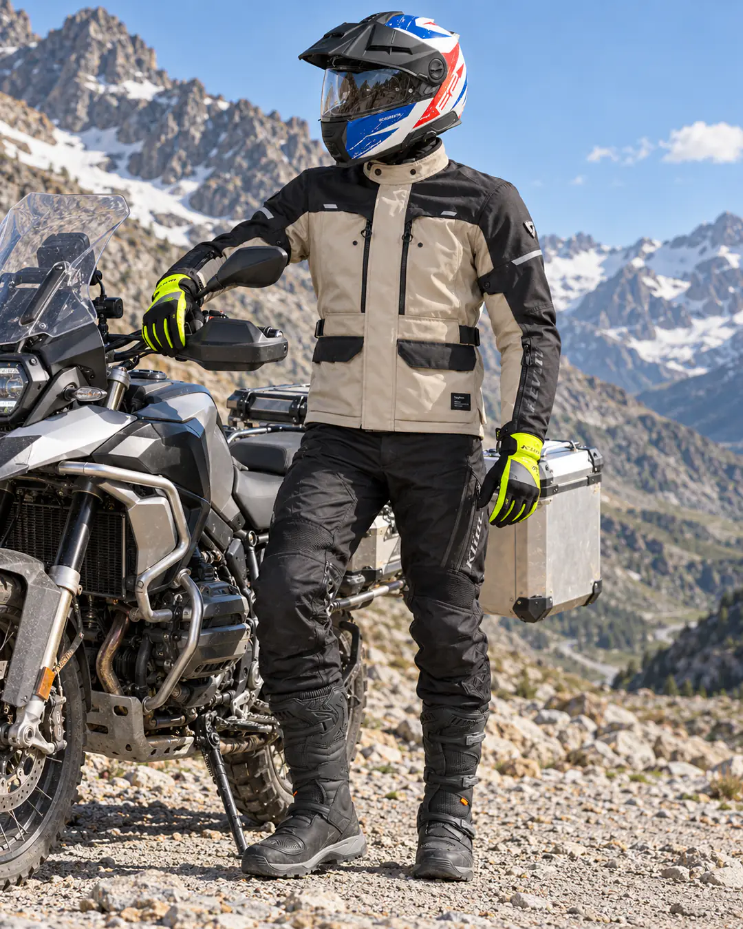 Motard près d’un gros trail à valises alu sur une piste de col de haute montagne enneigé, en casque Schuberth E2 Explorer bleu, blanc et rouge, veste Rev’it Outback 5 beige et noire, gants KLIM jaunes, pantalon KLIM Dakar et bottes Forma Adventure.